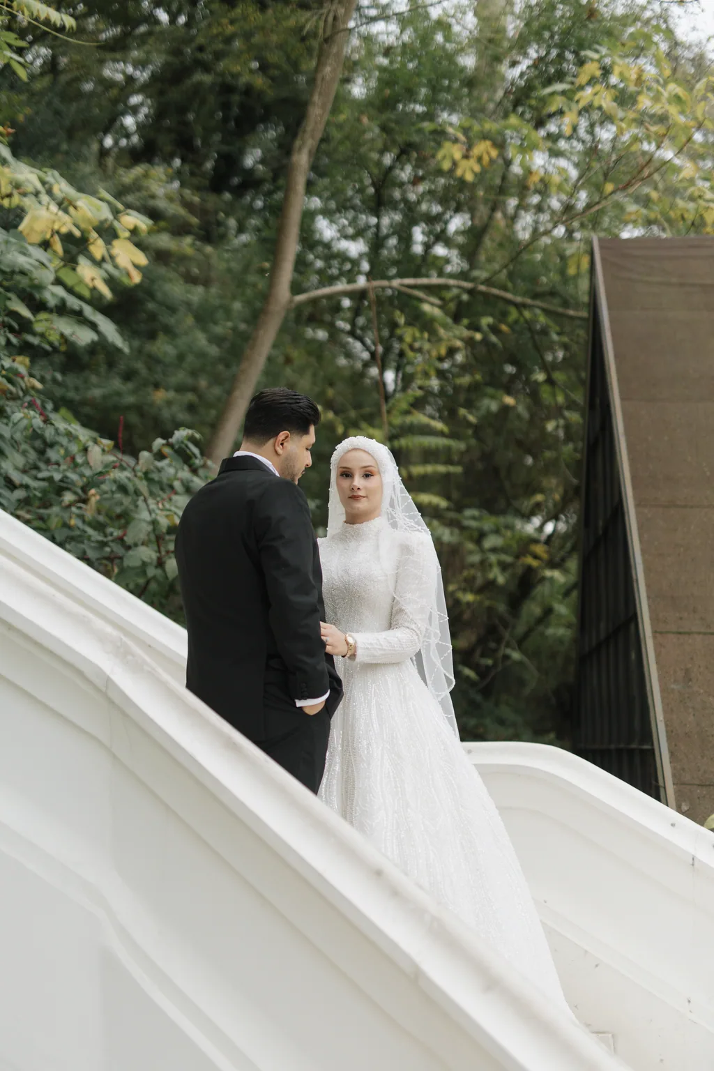 Gelin ve damat, ağaçların arasındaki beyaz merdivende yan yana duruyor.