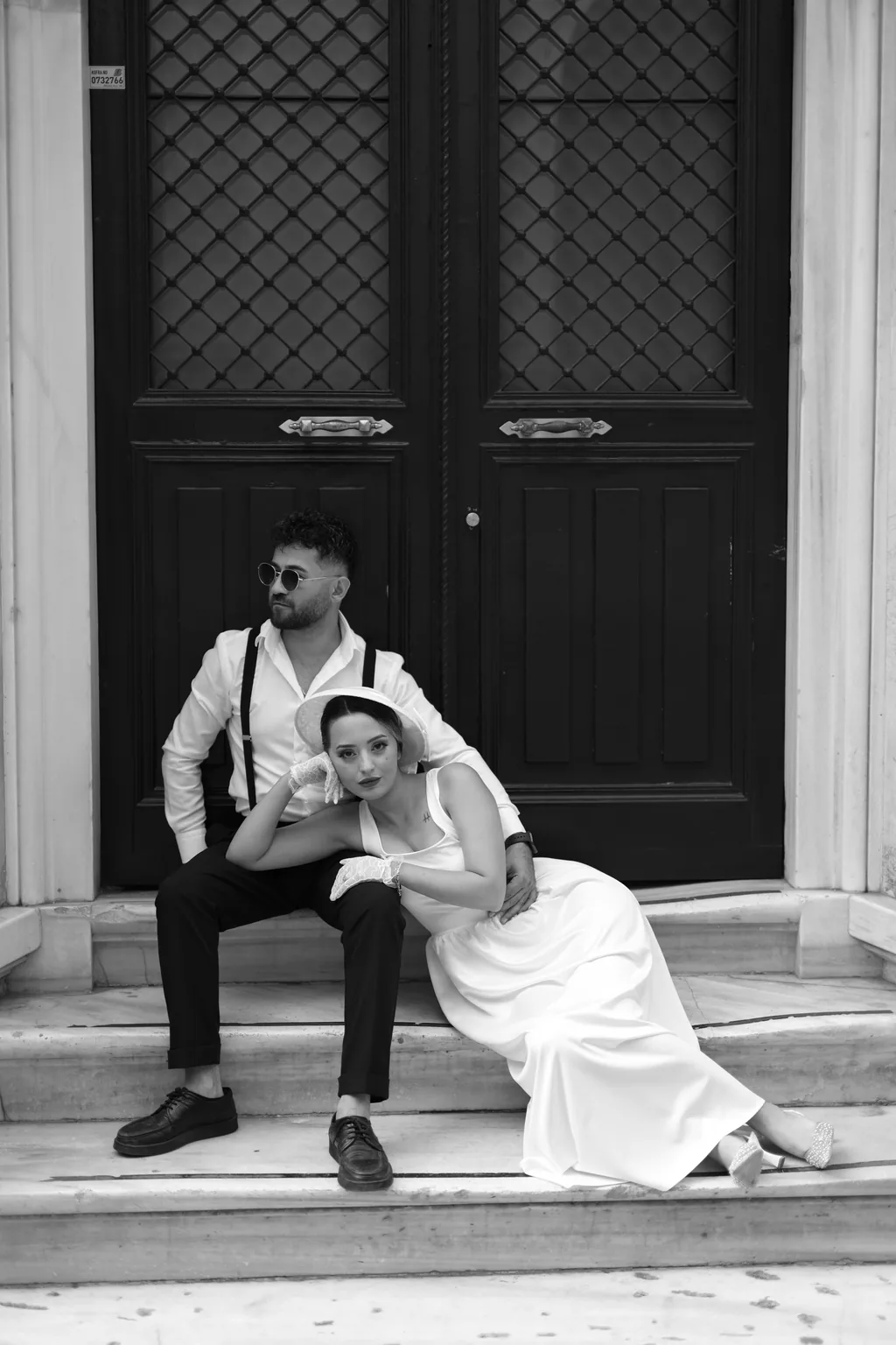 Siyah beyaz: damat siyah bir kapının önündeki basamaklarda oturuyor, gelin kucağına uzanmış.