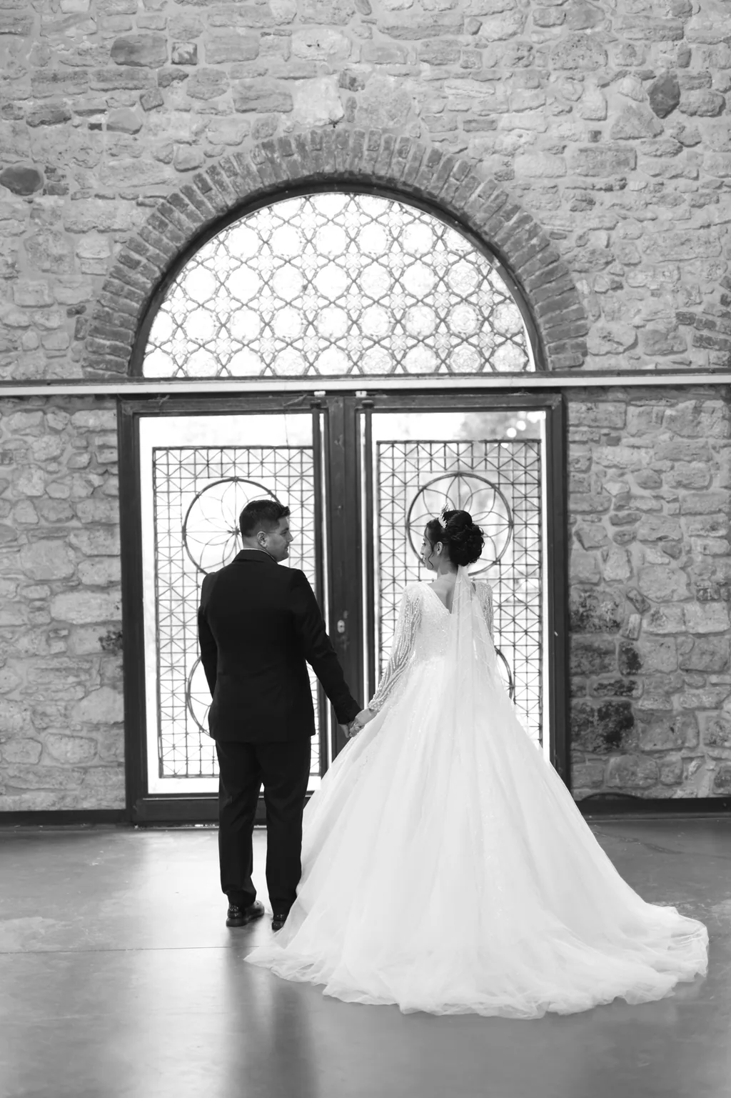 Siyah beyaz: gelin ve damat, taş duvardaki kemerli demir kapıya doğru yürüyor.