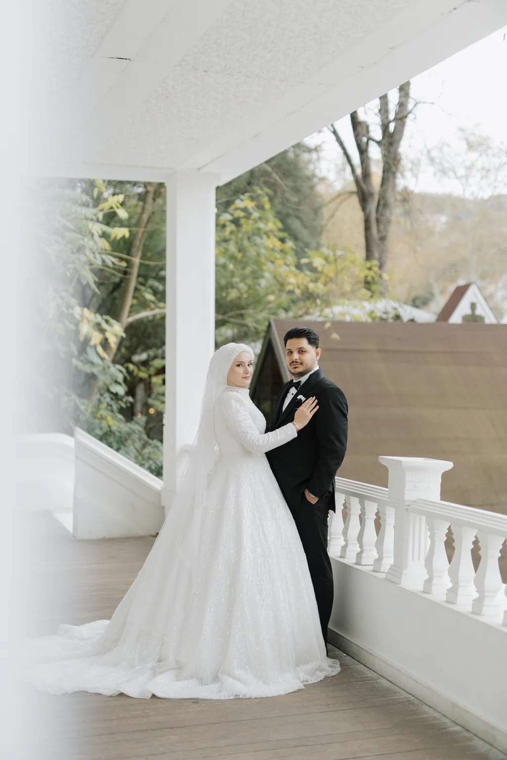 Duvaklı gelin ve damat, beyaz korkuluklu terasta yan yana.