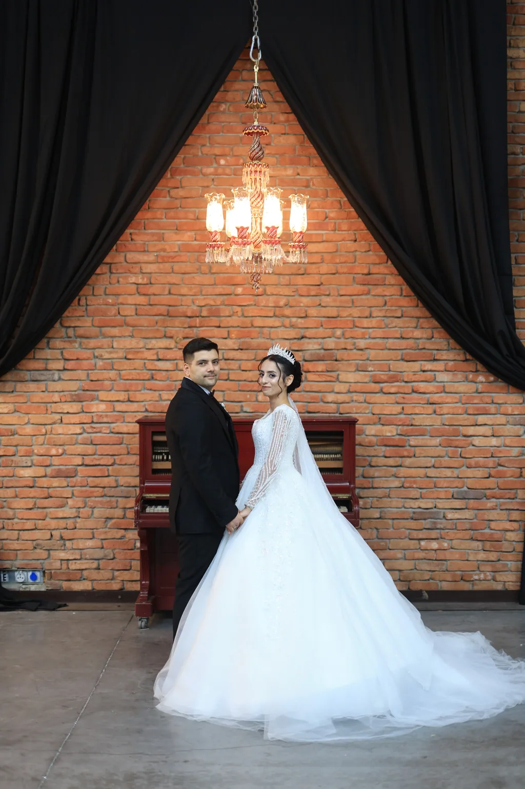 Gelin ve damat piyanonun önünde; arkada tuğla duvar, siyah perdeler ve avize.